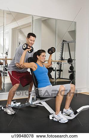 Stock Photo - Man assisting woman at gym with hand weights smiling.. Fotosearch
