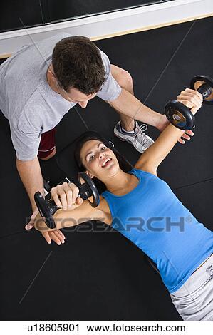 Stock Image - Man assisting woman lifting weights at gym. Fotosearch