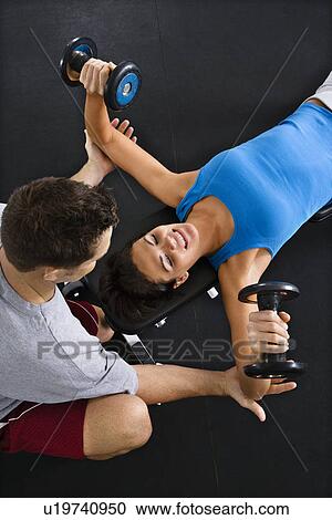 Stock Image - Man assisting woman lifting weights at gym.. Fotosearch