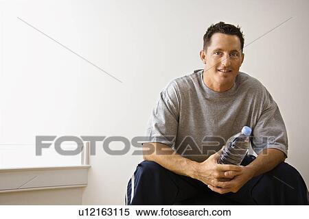 Stock Photography - Man holding water bottle sitting on balance ball at gym smiling.. Fotosearch