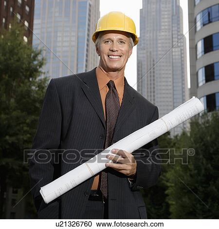 Stock Image - Middle aged businessman holding blueprints wearing hard hat . Fotosearch