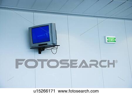 Stock Image - Monitor mounted to wall next to exit sign in airport. Fotosearch