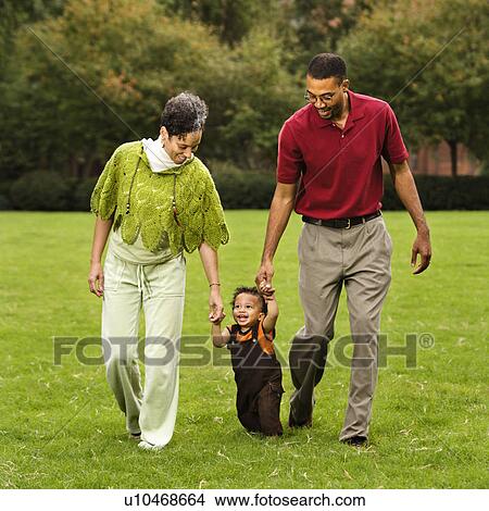 Picture - Mother and father helping toddler walk holding his hands in park.. Fotosearch