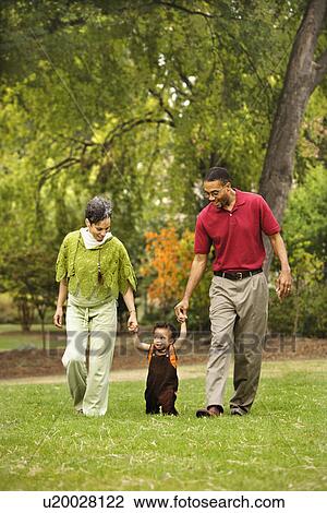 Stock Image - Mother and father helping toddler walk holding his hands in park.. Fotosearch
