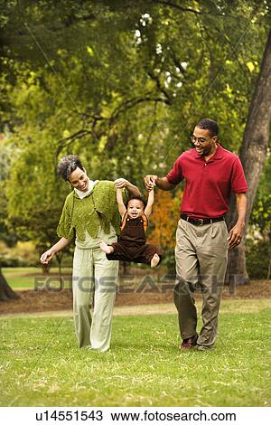 Stock Image - Mother and father swinging toddler holding his hands in park. Fotosearch