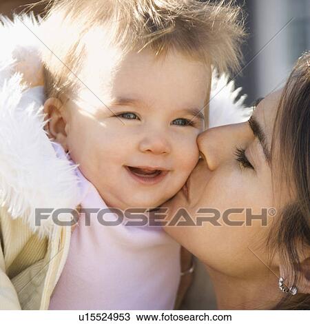 Stock Image - Mother holding and kissing smiling baby girl. Fotosearch
