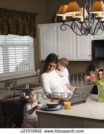 Mother holding baby and typing on laptop computer with girl eating breakfast in kitchen View Large Photo Image Stock Image - Mother holding baby and typing on laptop computer with girl eating breakfast in kitchen. Fotosearch
