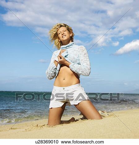Stock Image - Portrait of pretty young blond woman kneeling on Maui, Hawaii beach lifting up sweater.. Fotosearch