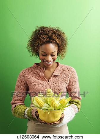 Picture - Portrait of pretty young woman standing against green background holding bowl full of ears of corn.. Fotosearch