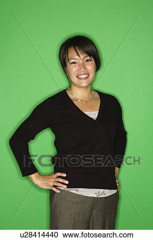 Stock Image - Portrait of smiling Asian woman standing with hands on hips against green background.. Fotosearch