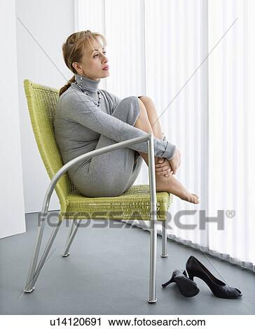 Stock Image - Pretty Caucasian woman sitting with legs curled up in chair looking away.. Fotosearch