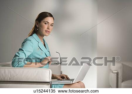 Stock Photography - Professional business woman sitting in modern office working on laptop computer holding eyeglasses and looking at viewer. Fotosearch