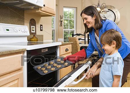Stock Photo - Side view of Hispanic mother and son putting cookies into oven.. Fotosearch