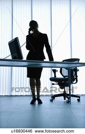 Stock Photograph - Silhouette of Caucasian businesswoman standing at computer desk on telephone.. Fotosearch