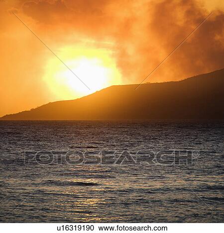 Stock Image - Sun setting behind mountain with ocean. Fotosearch