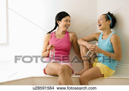 Picture - Two young women in fitness clothes holding water bottles sitting smiling and laughing and talking. Fotosearch