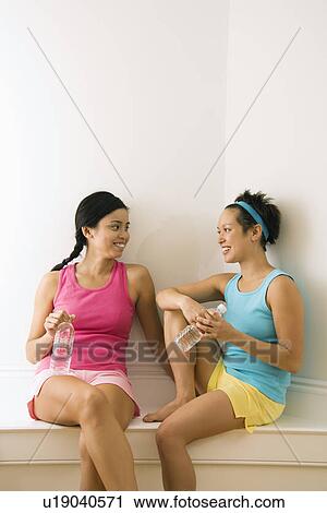 Two young women in fitness clothes holding water bottles sitting smiling and talking. View Large Photo Image Stock Image - Two young women in fitness clothes holding water bottles sitting smiling and talking.. Fotosearch