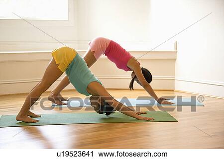 Stock Image - Two young women on yoga mats doing downward facing dog pose. Fotosearch