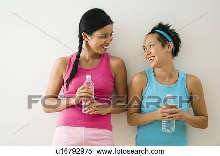 Stock Photography - Two young women standing in workout clothes talking and smiling holding bottled water.. Fotosearch
