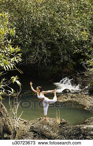 Stock Image - Woman doing yoga balancing on boulder by creek in Maui, Hawaii. Fotosearch