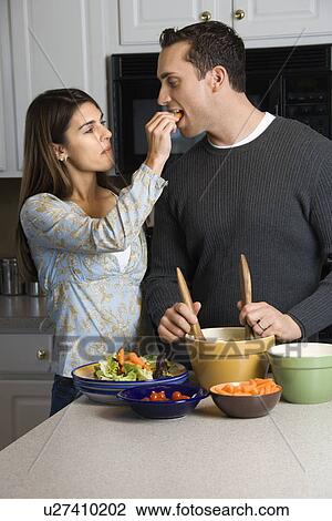 Stock Image - Woman feeding man at kitchen counter while he makes salad. Fotosearch