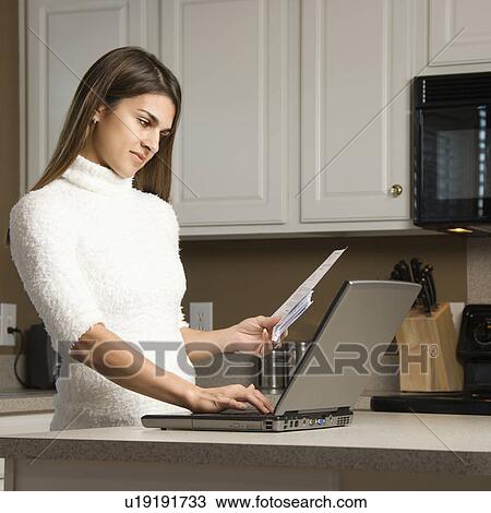 Stock Image - Woman in kitchen looking at laptop computer. Fotosearch