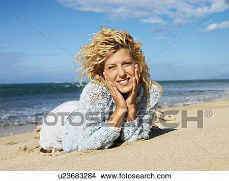 Picture - Woman lying in sand on beach with head resting on hands smiling. Fotosearch