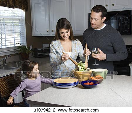 Stock Image - Woman making salad on kitchen counter with daughter and husband. Fotosearch