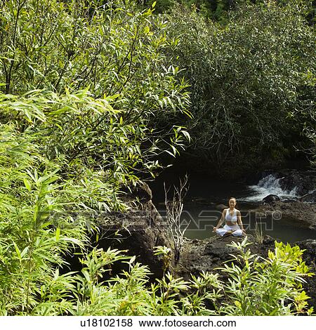 Stock Photo - Woman sitting on boulder by creek meditating. Fotosearch