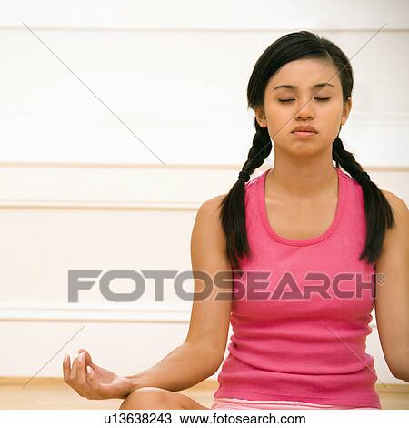 Stock Image - Woman sitting on floor meditating in yoga lotus pose with legs crossed and eyes closed. Fotosearch
