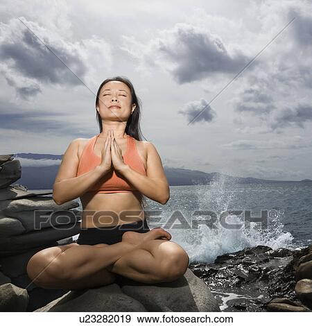Stock Photo - Woman sitting on rock by ocean in lotus pose with eyes closed. Fotosearch