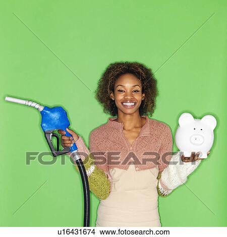 Picture - Woman standing holding gasoline pump nozzle and piggybank. Fotosearch