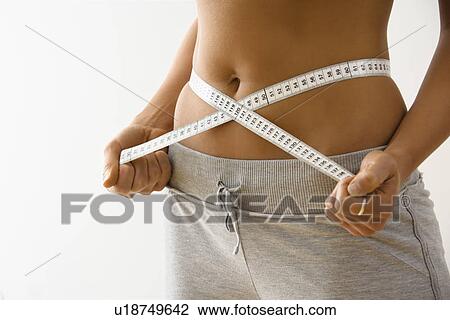 Stock Image - Woman standing pulling measuring tape around waist.. Fotosearch