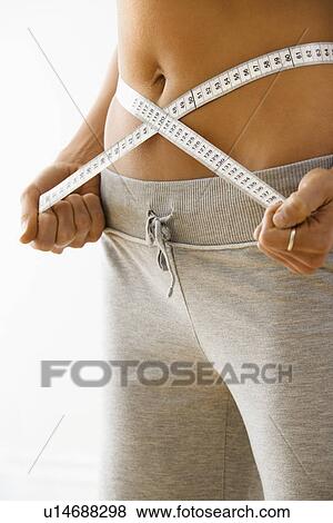 Stock Photo - Woman standing pulling measuring tape around waist.. Fotosearch
