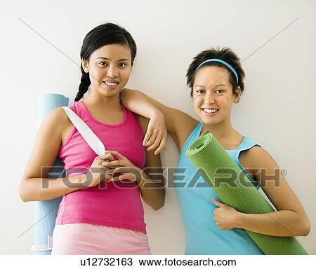 Stock Image - Women standing in workout clothes holding yoga mats and smiling. Fotosearch