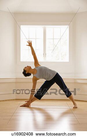 Stock Photo - Young woman doing yoga triangle pose indoors by sunlit window.. Fotosearch