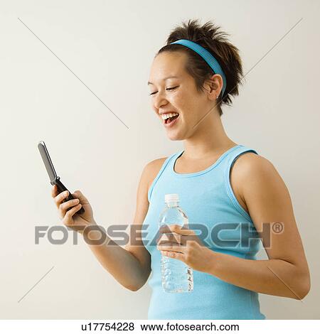 Young woman in fitness clothing holding bottled water and smiling at cellphone. View Large Photo Image Stock Photo - Young woman in fitness clothing holding bottled water and smiling at cellphone.. Fotosearch