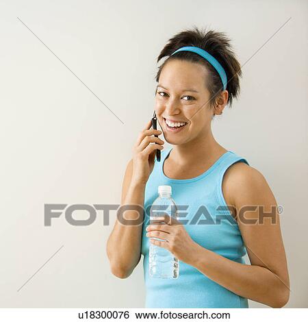 Stock Photograph - Young woman in fitness clothing holding bottled water talking on cellphone smiling.. Fotosearch