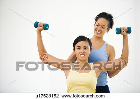 Stock Image - Young woman lifting hand weights while another woman helps position her arms. Fotosearch