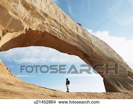 Picture - Hiker standing under arched rock. Fotosearch
