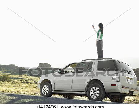 Stock Image - Woman using cell phone on roof of car, low angle view, Moab, Utah, USA. Fotosearch