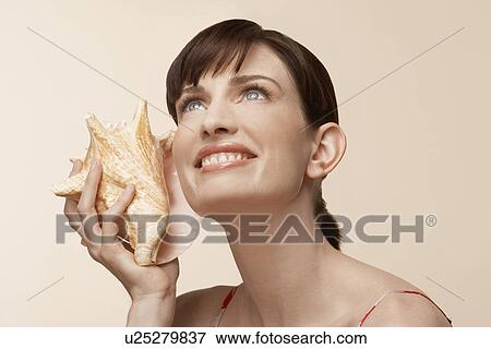 Stock Photo - Mid adult woman holding conch shell to ear. Fotosearch