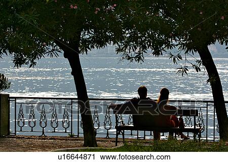 2 people sitting, talking, on bench at lakeside, Lake Como, Italy, silhouetted in evening sunlight, with shimmering water in background, and silhouette of trees framing them View Large Photo Image Stock Image - 2 people sitting, talking, on bench at lakeside, Lake Como, Italy, silhouetted in evening sunlight, with shimmering water in background, and silhouette of trees framing them. Fotosearch