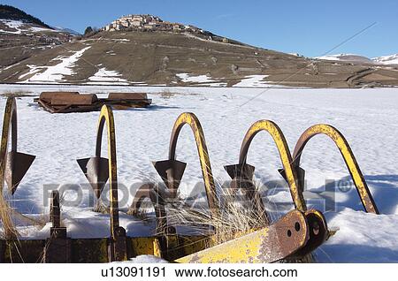 Stock Image - agricultural tools left for the winter in meadow of Piano Grande, Umbria, with town of Castellucio on hilltop beyond. Fotosearch