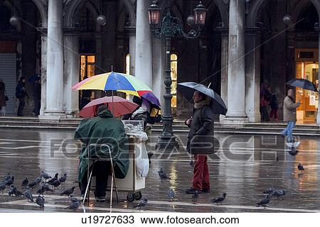 Bird food seller in St Marks Square under umbrella in rain, ornamental lamp and shopping arcade in background, pigeons, reflections in wet pavement View Large Photo Image Stock Image - Bird food seller in St Marks Square under umbrella in rain, ornamental lamp and shopping arcade in background, pigeons, reflections in wet pavement. Fotosearch