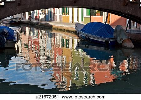 Stock Image - Burano canal with moored boats and reflections of brightly coloured buildings. Fotosearch