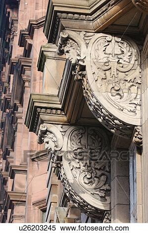 Stock Photography - Detail of facade decorations over main entrance with ornate stone scrollwork and faces, mock Renaissance style, and red fronted facade in background, Glasgow, Scotland. Fotosearch