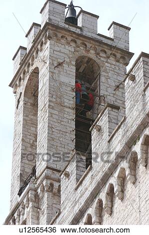 Stock Photograph - Detail of square bell tower of Palazzo dei Consoli, Gubbio, with men ringing the bells by swinging on them during May Day festivity. Fotosearch