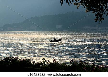 Stock Image - Dinghy in motion on Lake Como, Italy, with trees silhouetted in foreground, in evening light, water shimmering and mountains in background. Fotosearch
