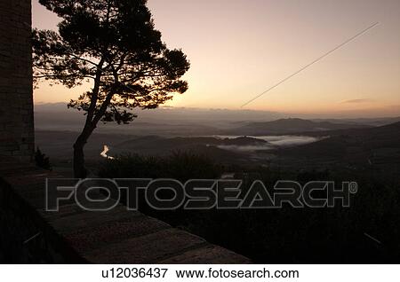 Stock Photo - Evergreen tree silhouetted beside old stone town wall at sunrise, with view over Tiber valley in misty dawn of winter, river winding through valley. Fotosearch